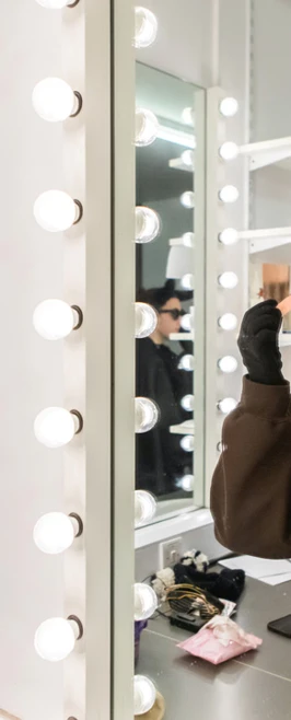 Student applying theatrical makeup while looking into a mirror with bright makeup lights Student applying theatrical makeup while looking into a mirror with bright makeup lights